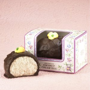 16-oz Dark Chocolate Coconut Cream Eggs with edible yellow decorative flowers on top. One (1) Egg cut in half to reveal white inside. Other egg whole in package with clear window to show outside of egg. Pictured on light purple backdrop.