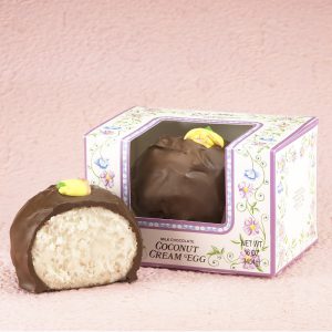 16-oz Milk Chocolate Coconut Cream Eggs with edible yellow decorative flowers on top. One (1) Egg cut in half to reveal white inside. Other egg whole in package with clear window to show outside of egg. Pictured on light purple backdrop.