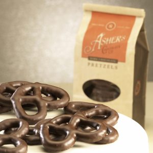 Dark Chocolate Pretzel Coffee Bag in background with whole three (3) ring pretzels on white plate in front. Coffee Bag is brown with orange details. Clear plastic circular window on front displays Dark Chocolate Pretzels inside.