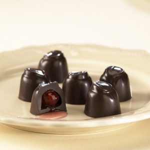 Dark Chocolate Cordial Cherries sit on white plate. One (1) cordial is cut open to reveal a sweet, red cherry inside.