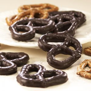 Dark Chocolate three (3) ring Pretzels scattered on white plate. Size, shape, and texture are shown compared to non-chocolate coated pretzels in the background.