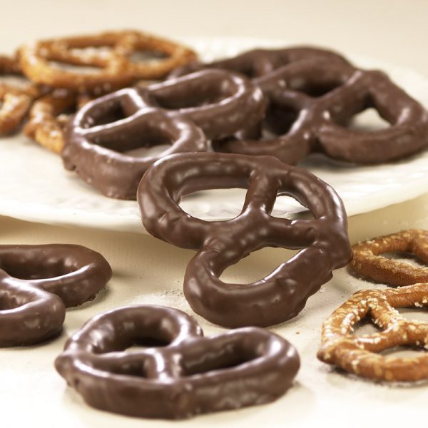 Milk Chocolate three (3) ring Pretzels scattered on white plate. Size, shape, and texture are shown compared to non-chocolate coated pretzels in the background.