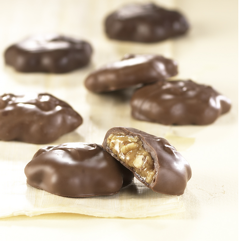 Milk Chocolate Pecan Caramel Patties are scattered on a white cloth. One (1) Pecan Caramel Pattie is broken open to reveal the caramel and nuts on the inside of the candy.