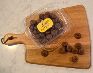 Milk Chocolate Wilbur Bubs Fresh Pack displayed on a wooden board. Some milk chocolate bubs are scattered outside of fresh pack, displaying shape, and color. Rest of bubs fill fresh pack with gold and brown Asher's sticker labeling the lid.